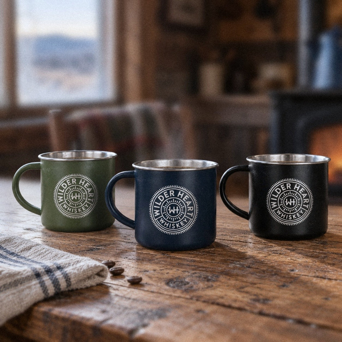 Wilder Heart camp mugs in Green, Navy and Black on a cabin kitchen table at dawn
