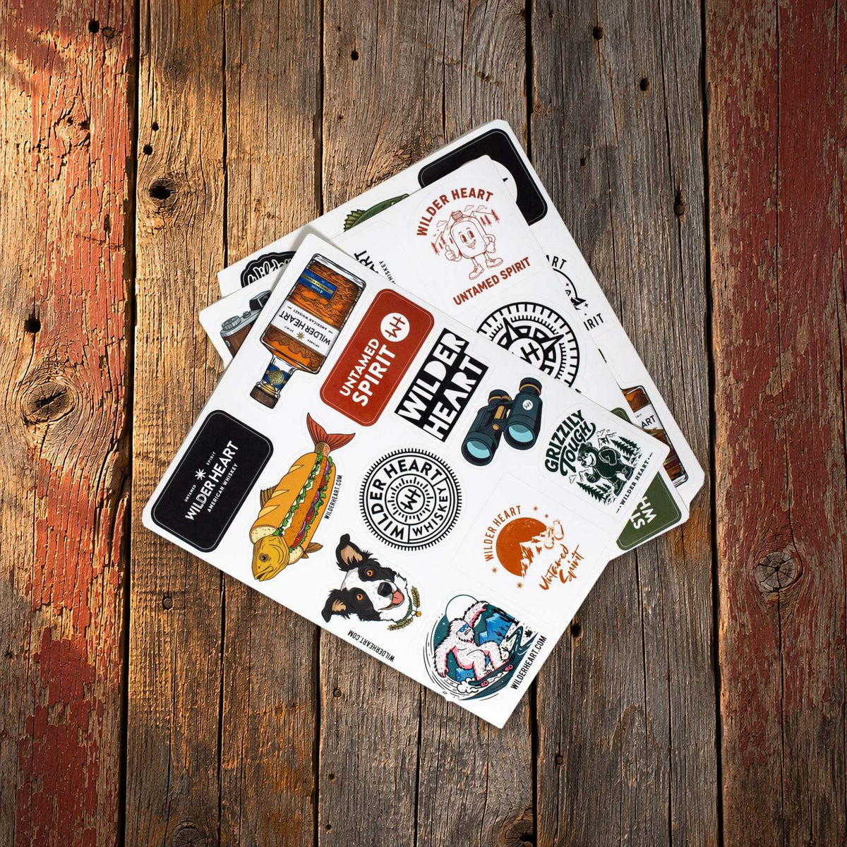 All three Wilder Heart sticker sheets fanned out on a weathered barn-board table: Trail Dog, Big Bass and Compass