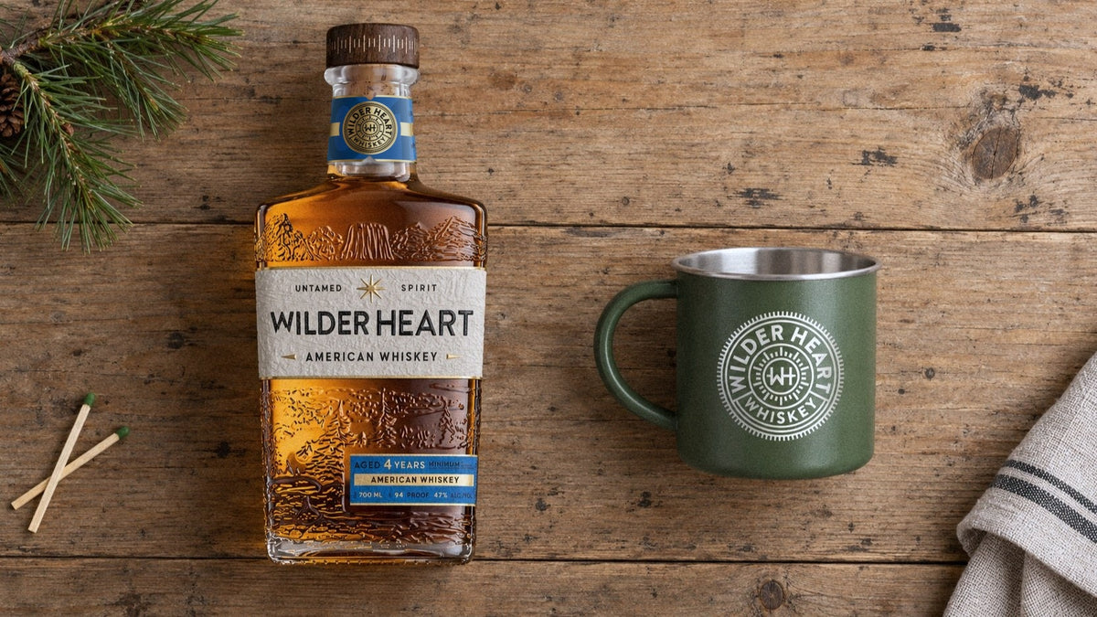 The Fireside Kit laid out on a pine table: Wilder Heart Whiskey and the camp mug