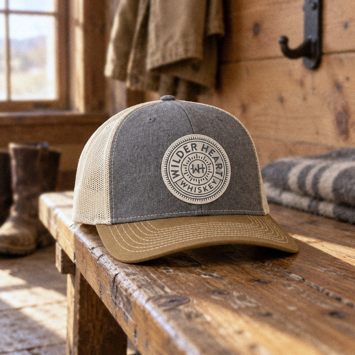 The Wilder Heart Richardson 112 trucker hat in heather gray with a cream mesh back, tan bill and cream PVC medallion patch, on a worn pine bench in a ranch-house mudroom