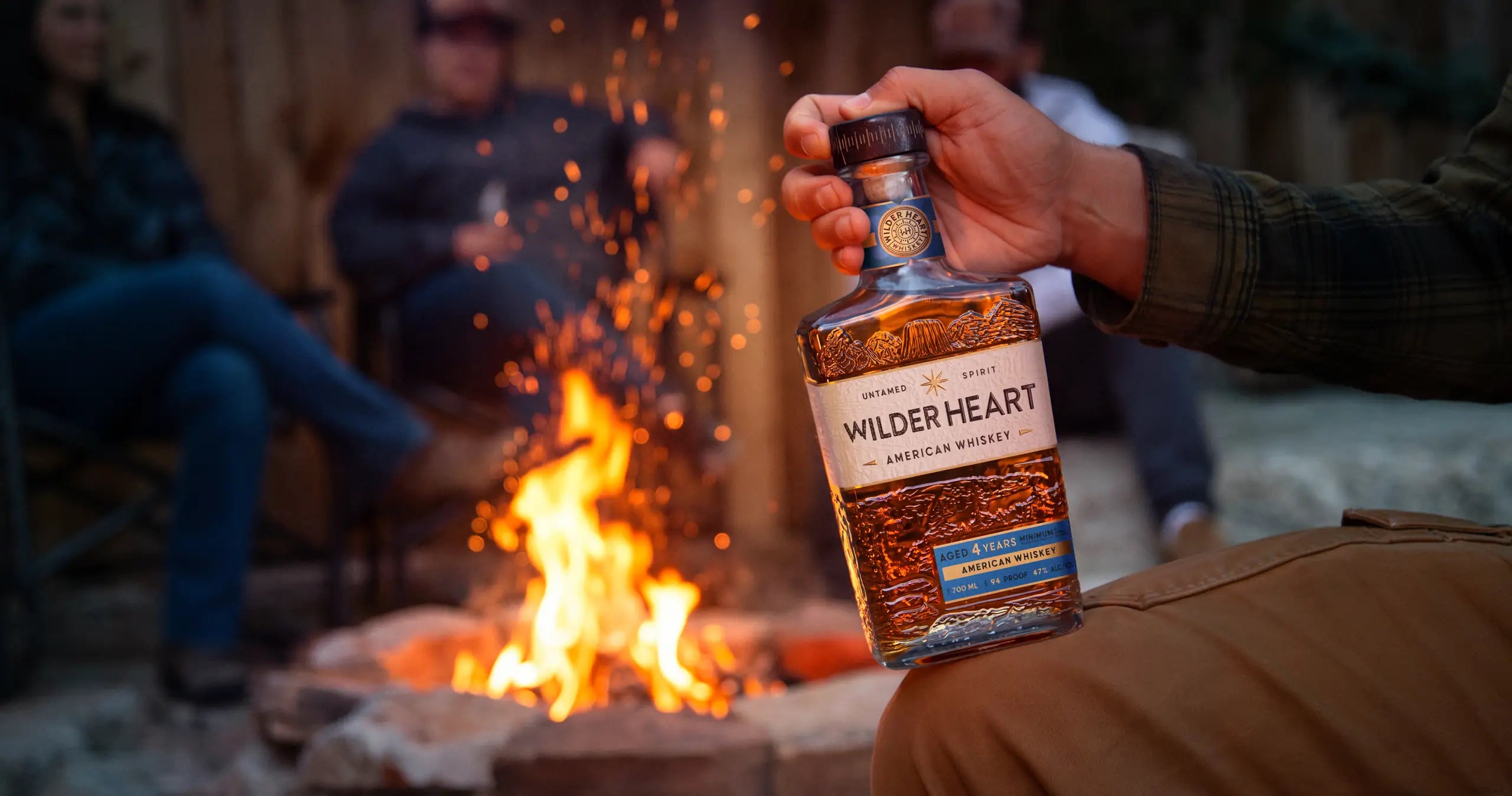 A hand holding a bottle of Wilder Heart by the campfire