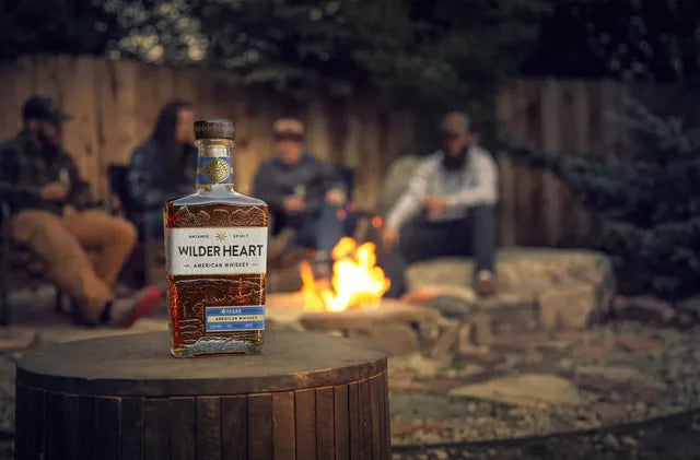 Friends around a campfire at night with a bottle of Wilder Heart on a stump