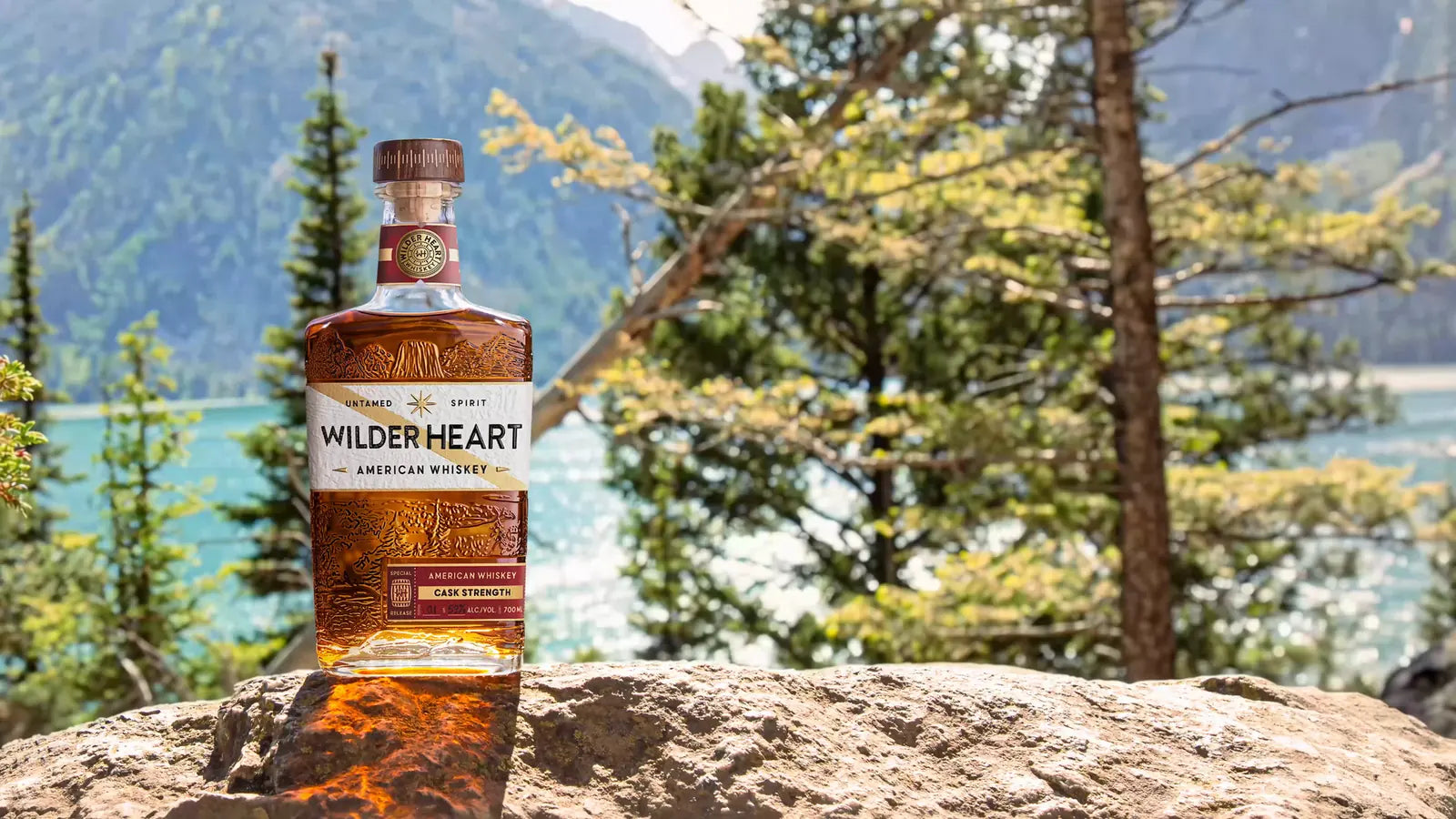 A bottle of Wilder Heart Cask Strength Whiskey on a rock among the pines, a mountain lake behind it