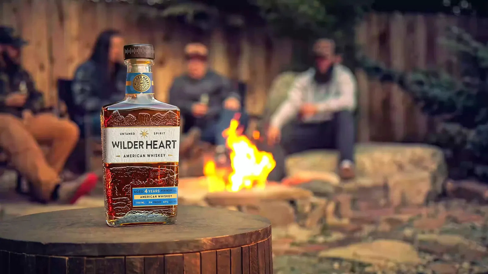A bottle of Wilder Heart American Whiskey on a table by the campfire, friends gathered behind it