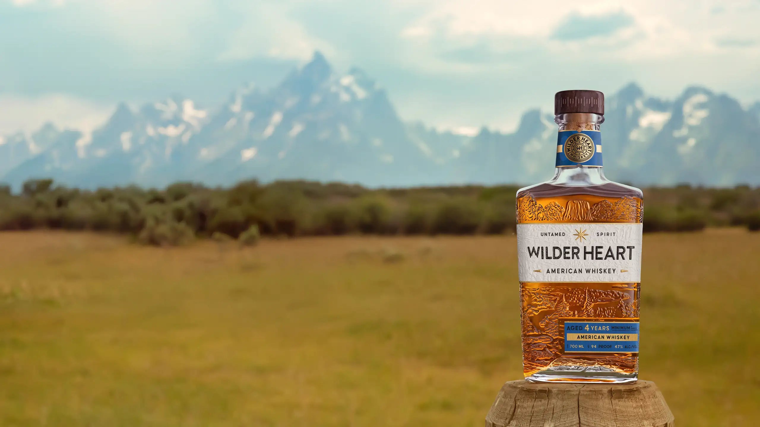 A bottle of Wilder Heart on a fence post in a meadow below the Tetons